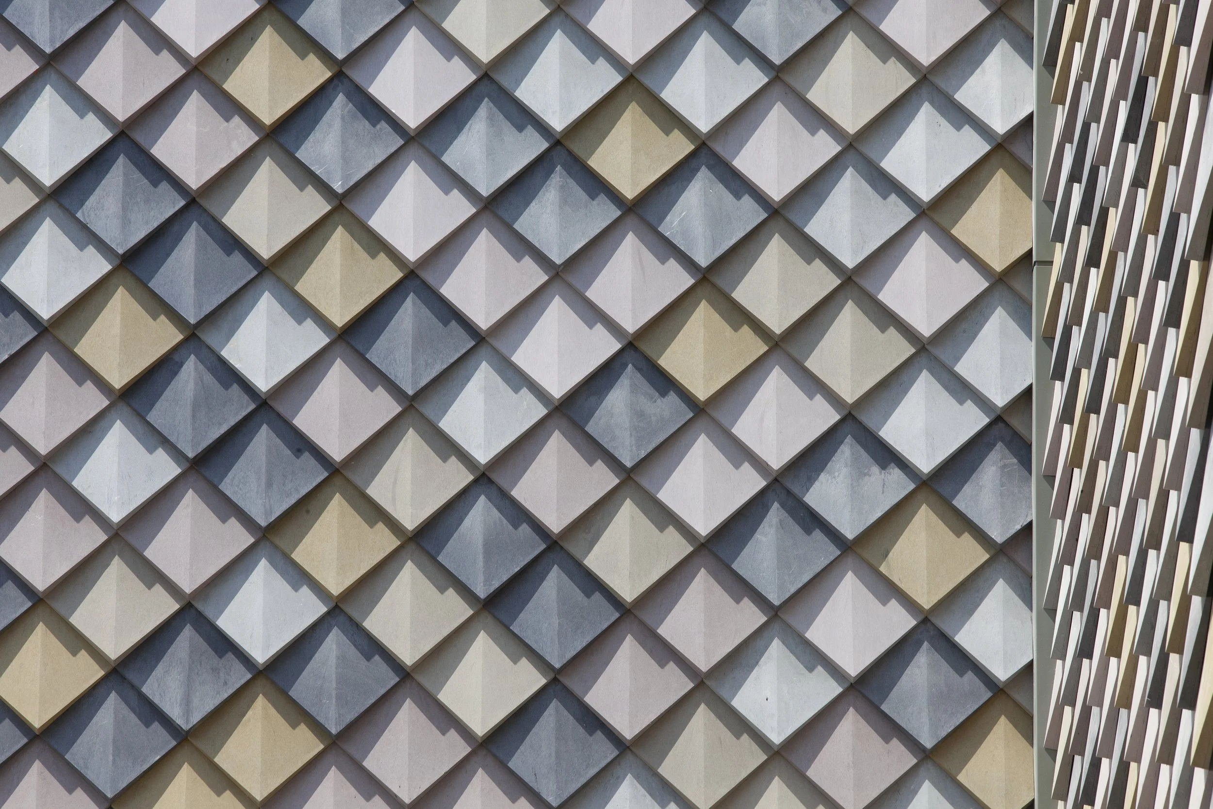 Close-up of a building's exterior wall decorated with light-colored, diamond-shaped tiles arranged in a repeating pattern, with a section of a window or decorative element on the right side. Howest in Brugge