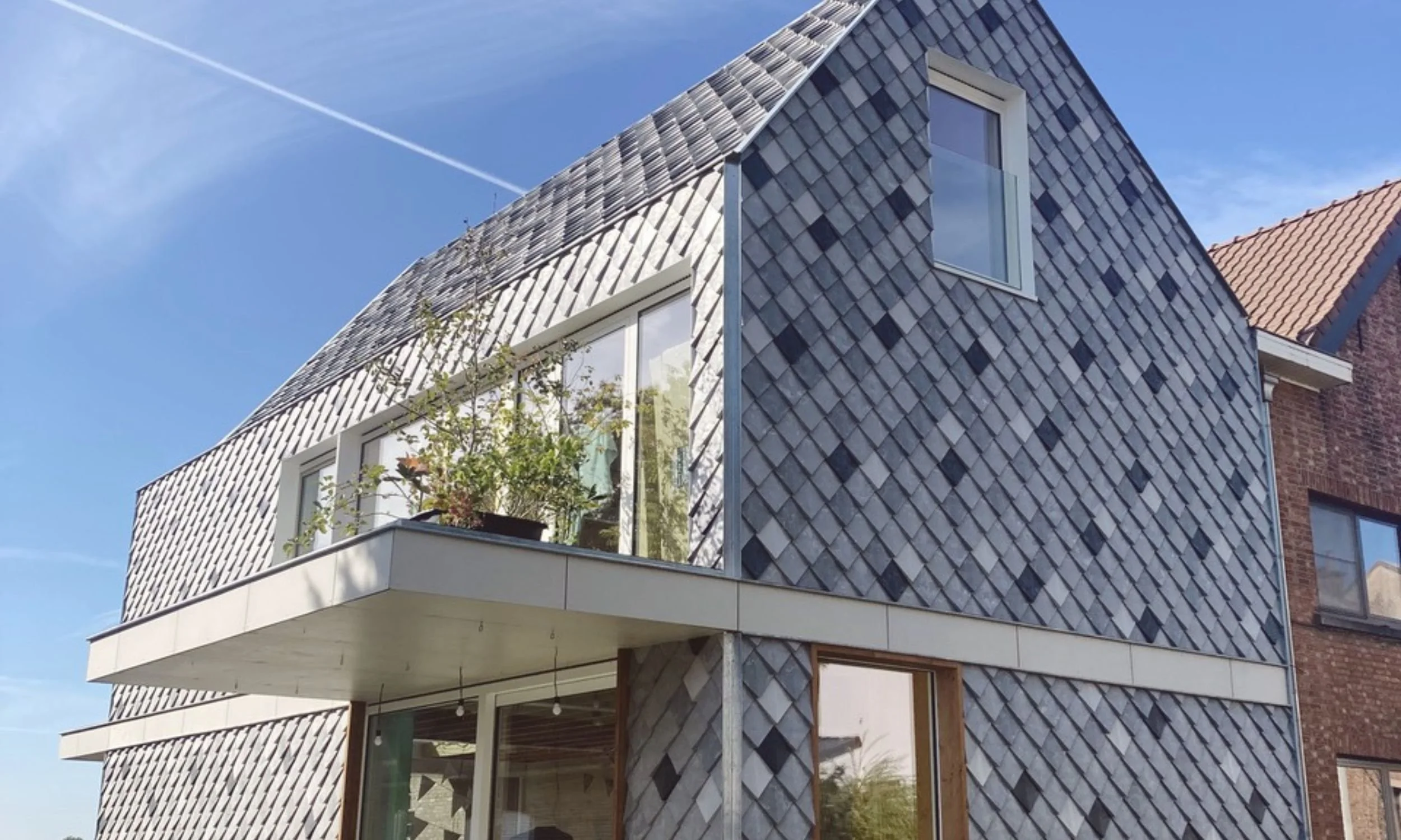 Modern residential building with a unique exterior design featuring a mix of slate tiles and large windows, with a balcony and some plants outside, under a clear blue sky with a contrail.