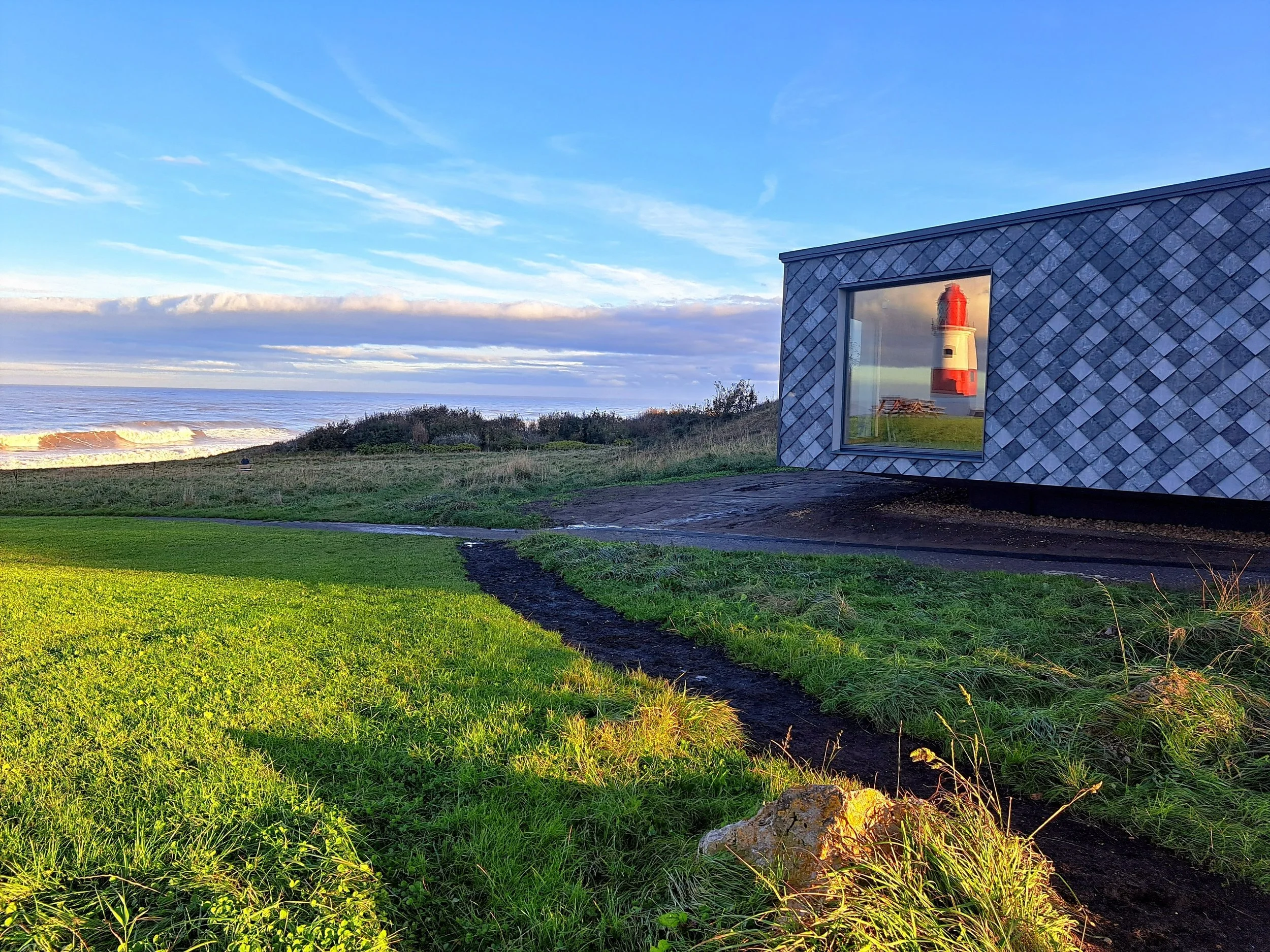 A landscape view of the beach at sunset. The sky is blue with clouds, and the ocean waves are visible. On the right, a modern building with a window reflecting a lighthouse and picnic tables is seen. The foreground has green grass and a dirt pathway.