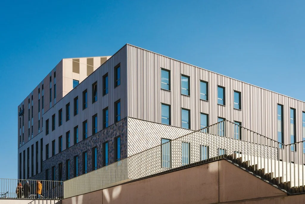 Modern multi-story building with metal and glass exterior, blue sky, and a few people walking on a balcony in the foreground.