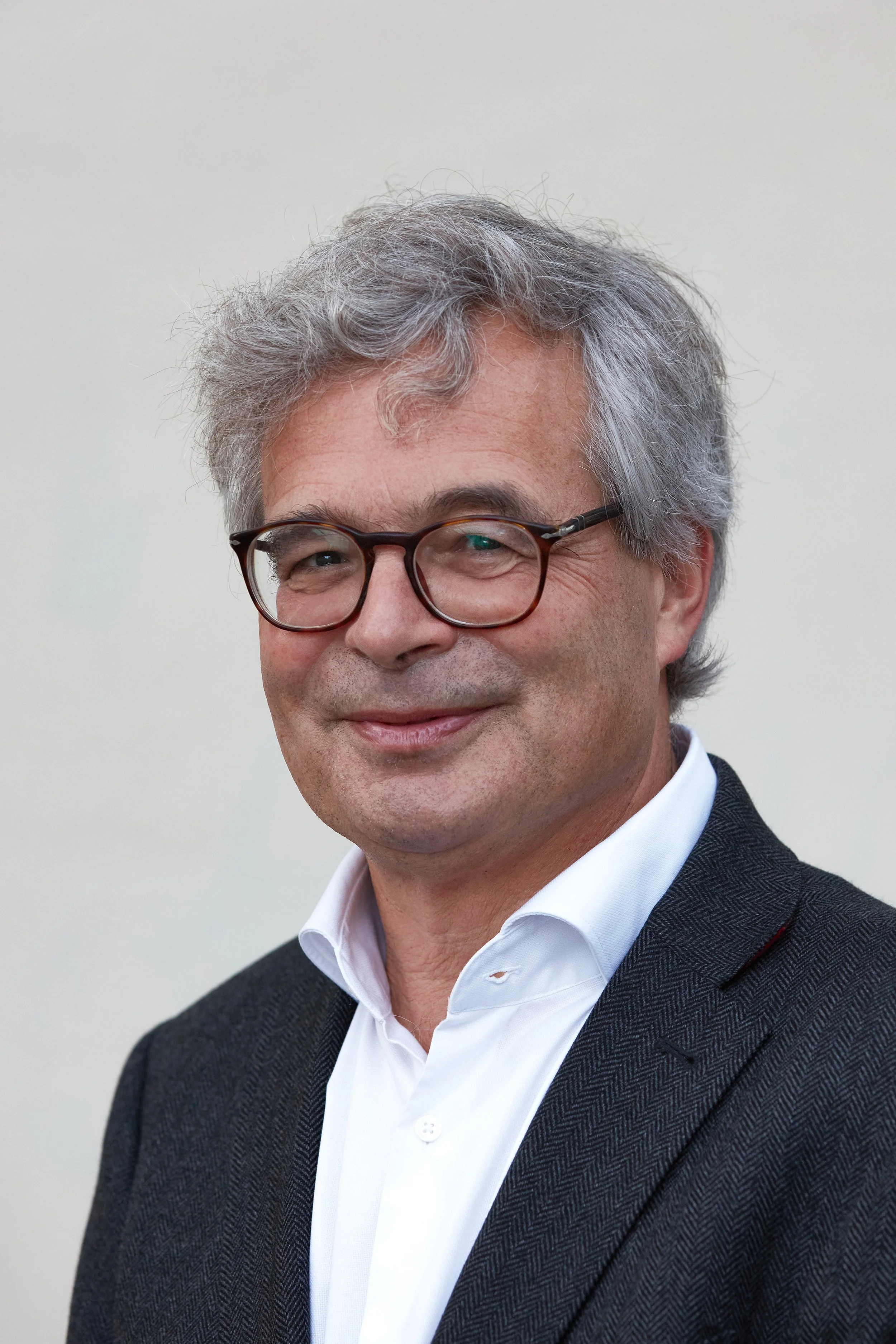 Close-up of a middle-aged man with gray, wavy hair, wearing glasses, a white dress shirt, and a dark blazer, standing in front of a plain light-colored wall.