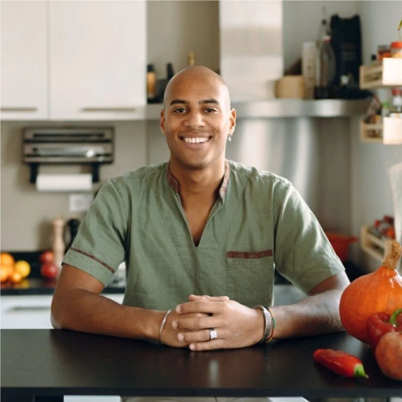 A smiling man sitting at a kitchen counter with pumpkins and a pepper on it.