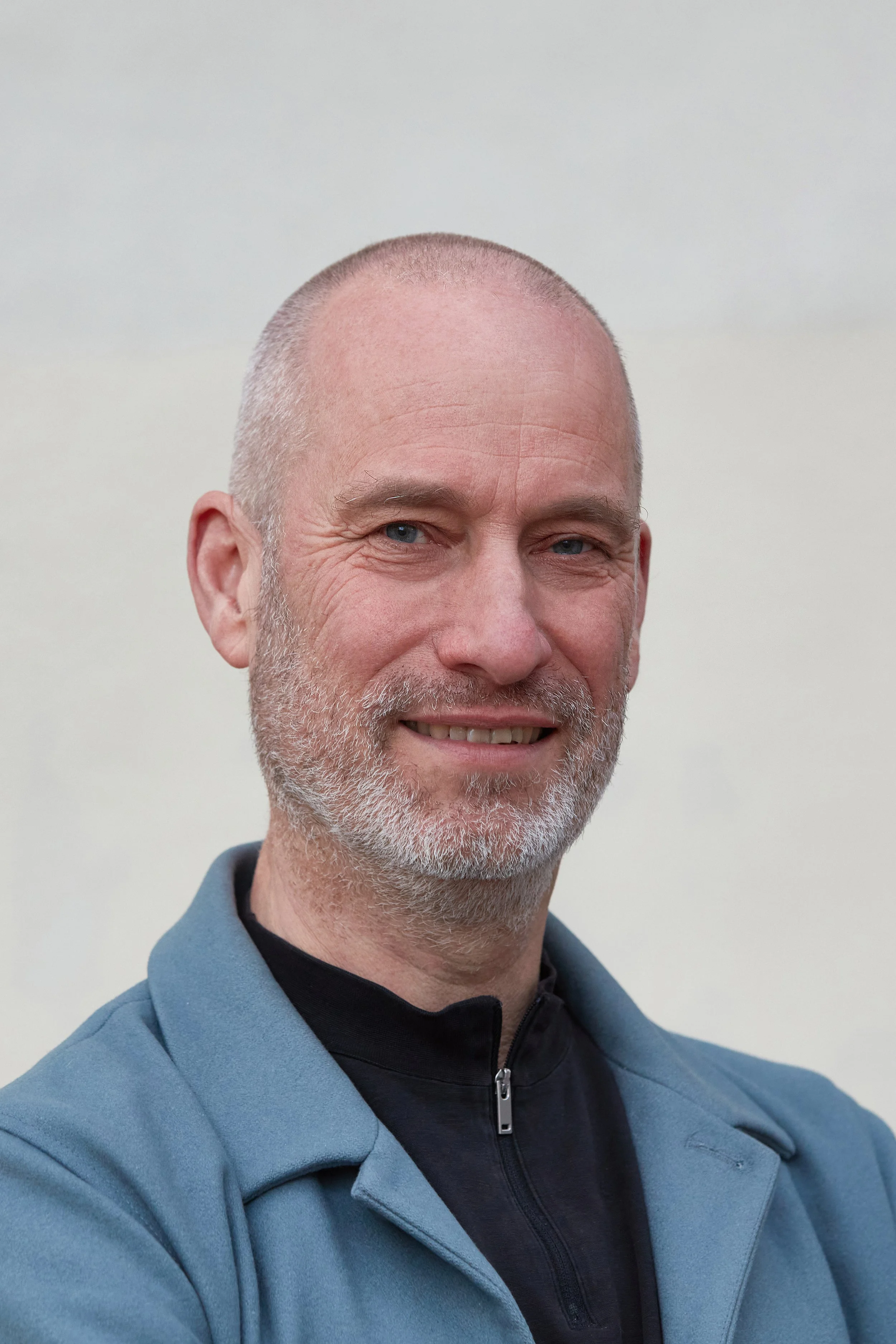 A middle-aged man with a shaved head and gray beard smiling, wearing a light blue jacket over a black zip-up shirt, standing against a plain white background.