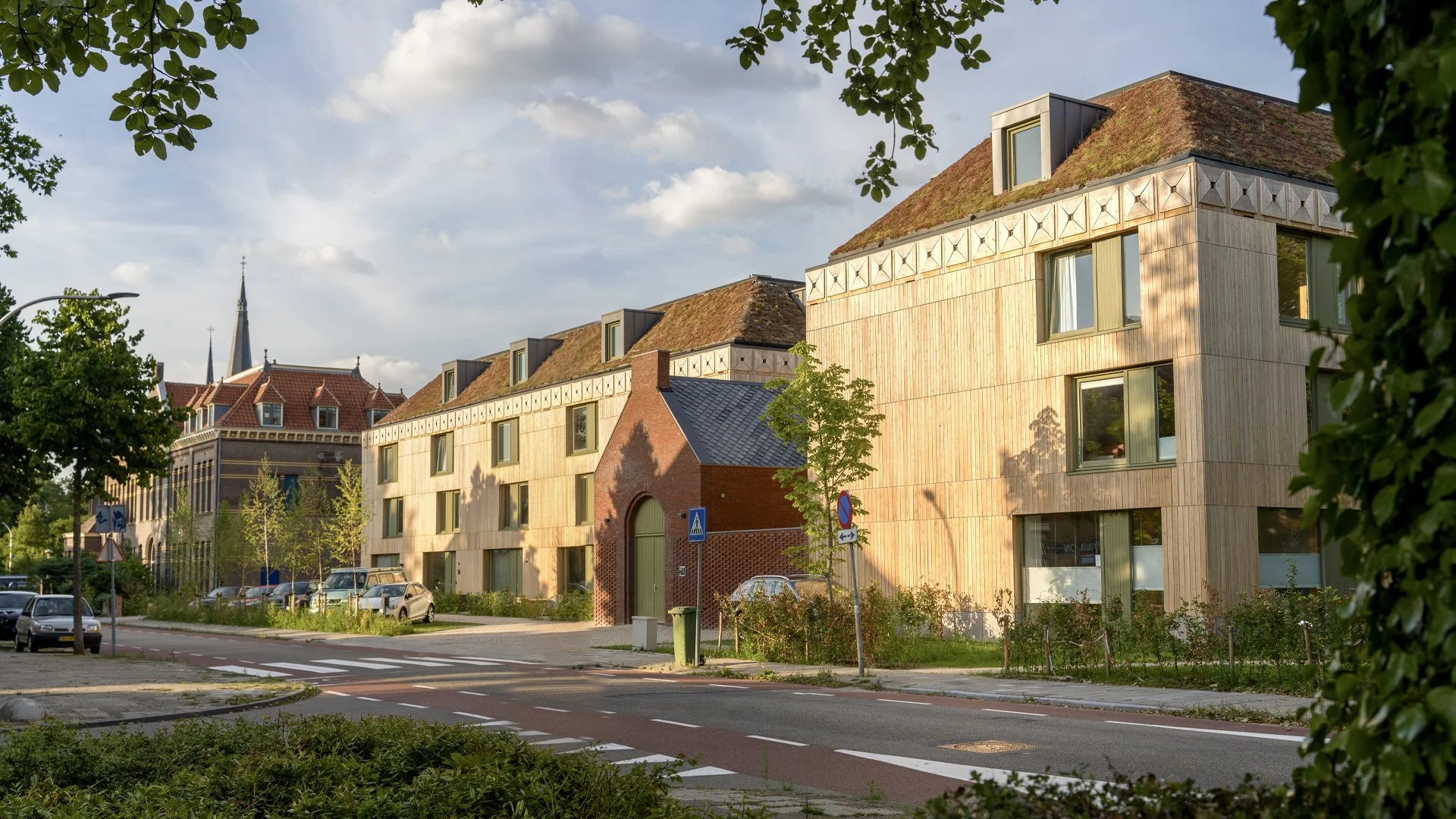 Modern wooden apartment building with in the background a traditional church steeple, trees and parked cars along the street in late afternoon light.