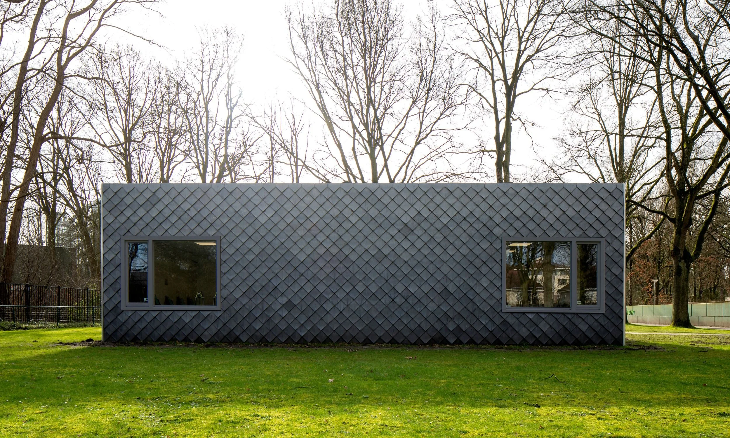 A small modern building with gray diamond-patterned siding and two large windows, set on a grassy area with a background of leafless trees and a partly cloudy sky.