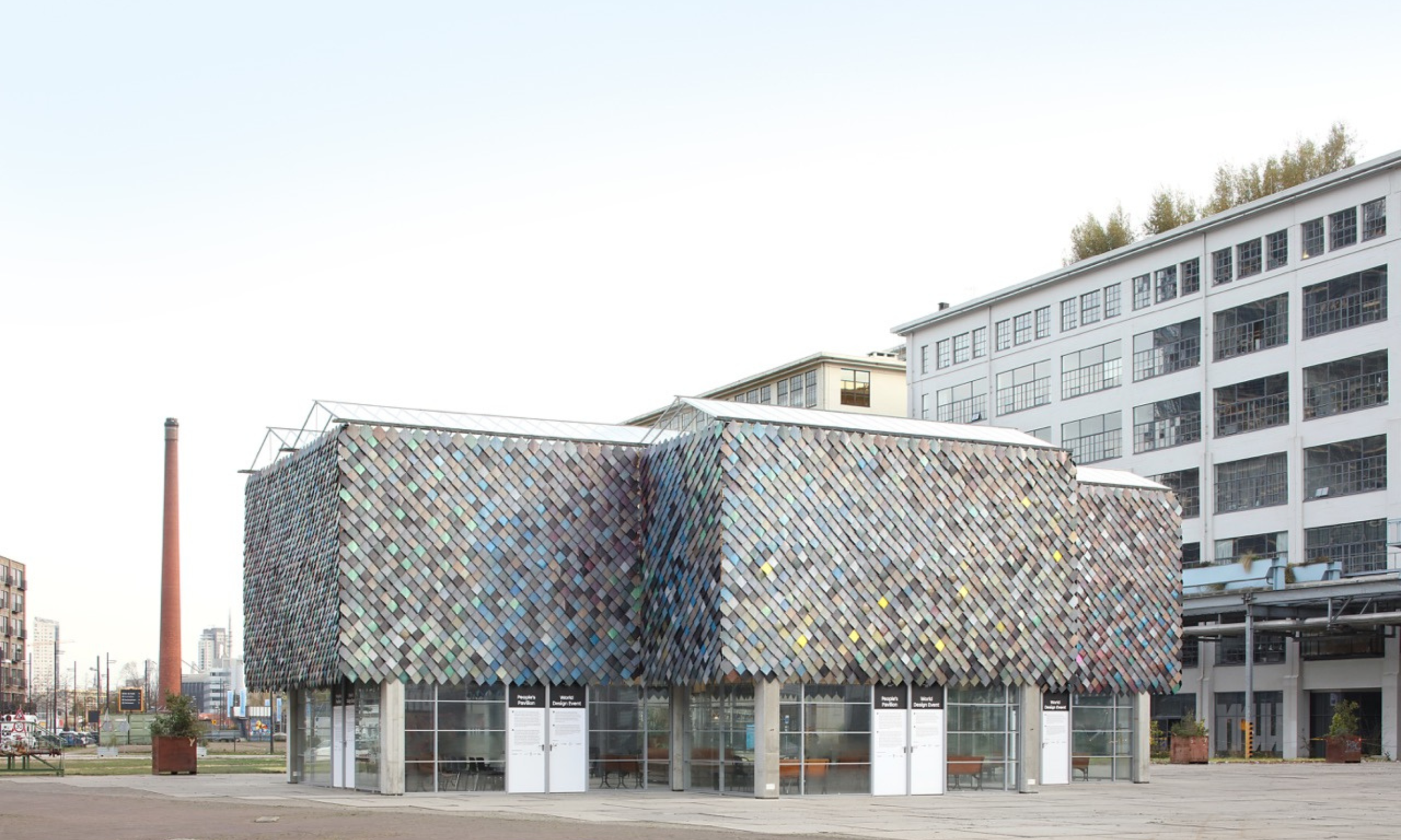 Deze foto biedt een panoramisch zicht op The People's Pavilion, een toonbeeld van circulaire innovatie en duurzaamheid. Met zijn gevel die schittert in een rijk palet van kleuren, allemaal afkomstig van gerecyclede materialen, en zijn dak en ramen di